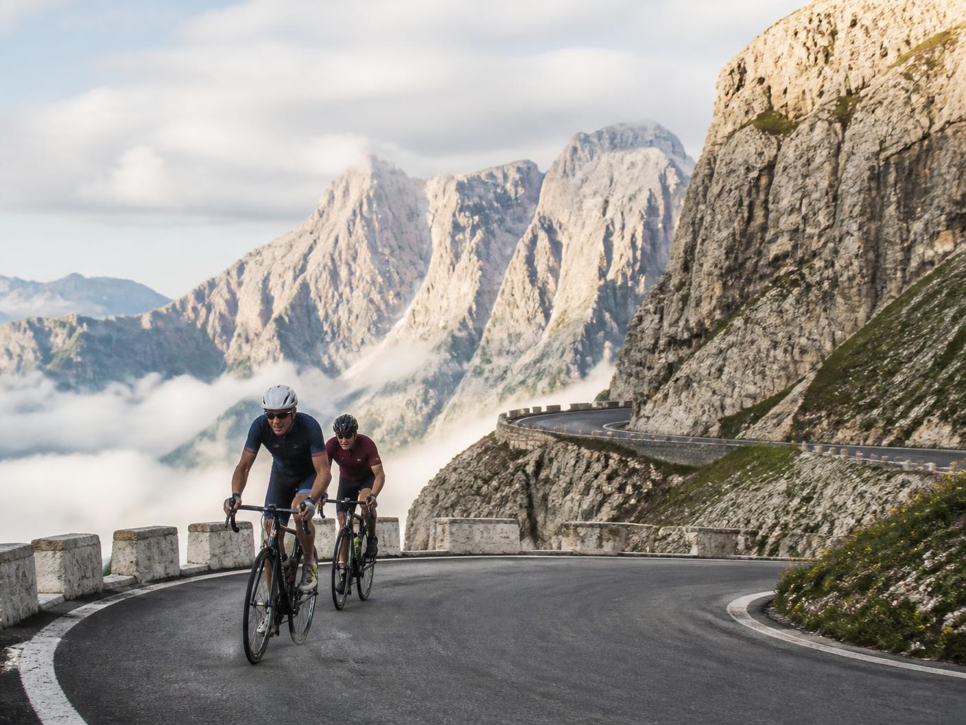 Two experienced cyclists climbing a dramatic alpine hairpin road in the Dolomites with limestone peaks and morning mist below.